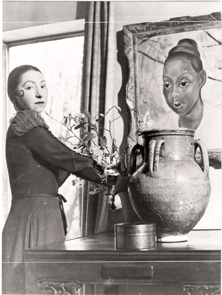 Eine Frau steht links nebe einer großen Vase und blickt in die Kamera. Rechts im Bild ist ein großes Gemälde mit einem Frauenkopf abgebildet.