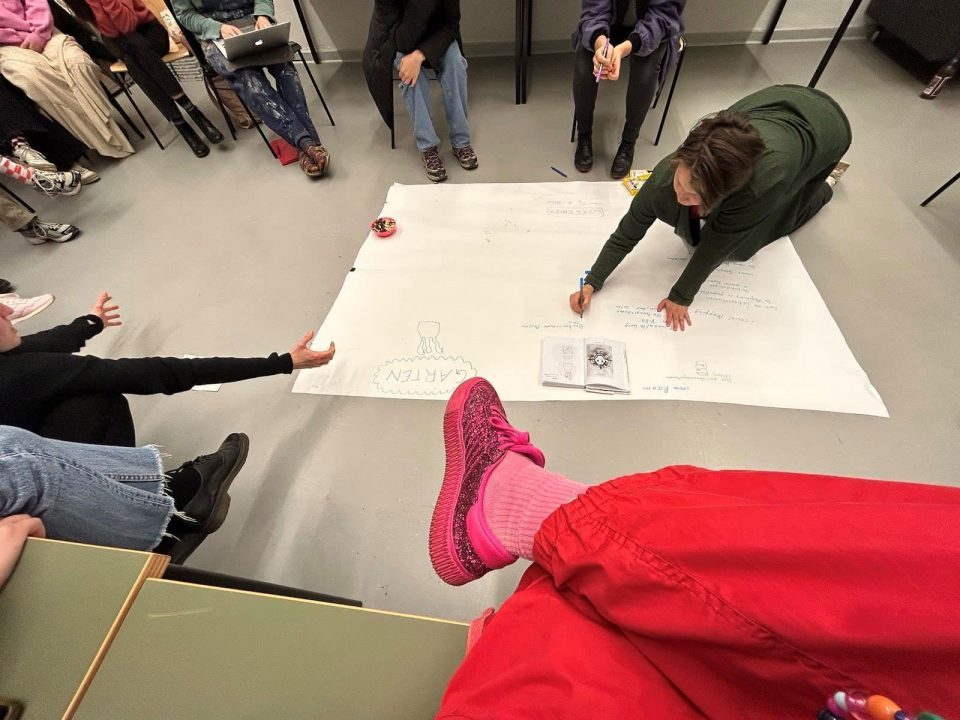Eine Studierende schreibt knieend ihre Gedanken auf einen großen weißen auf dem Boden ausgebreiteten Papierbogen. Sie wird umrundet von ihren StudienkollegInnen.