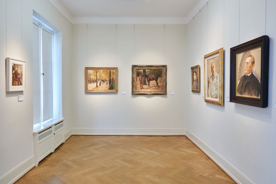 In einem Ausstellungsraum hängen sechs Gemälde an der Wand. Die meisten zeigen Personen. Links zwischen den Fenstern hängt das Selbstbildnis von Max Liebermann.