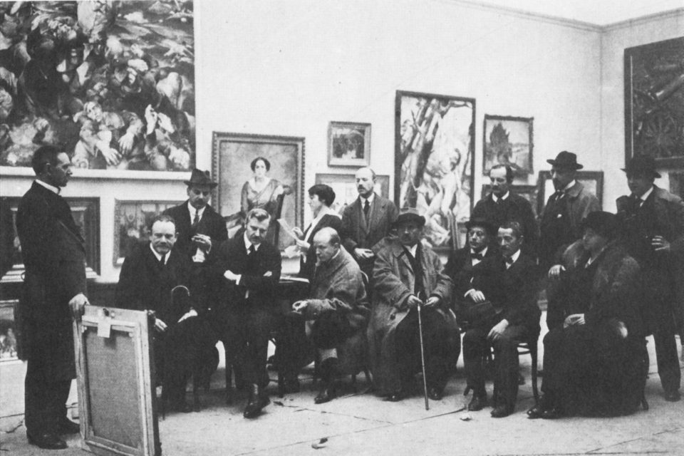 Eine Runde von Herren und Damen sitzt in einem Ausstellungssaal und betrachtet gemeinsam ein Gemälde, hinter ihnen hängen weitere Gemälde an der Wand