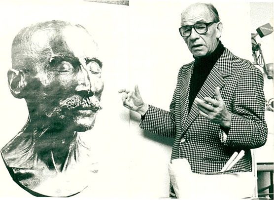 Der Bildhauer Arno Breker mit Brille und im karierten Sakko steht neben der Totenmaske, die er von Max Liebermann abgenommen hat. Hier ist ein Bronzeabguss gezeigt. 