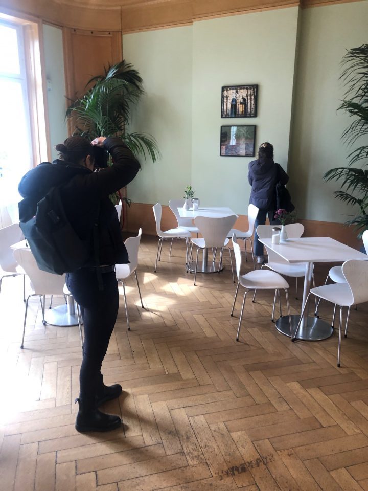 Zwei junge Kunststudierende sind von hinten zu sehen. Die eine steht hinter der anderen und fotografiert sie. Das Foto ist im Café der Liebermann-Villa aufgenommen, in den Ecken stehen große grüne Palmen und weiße Cafétische mit Stühlen.