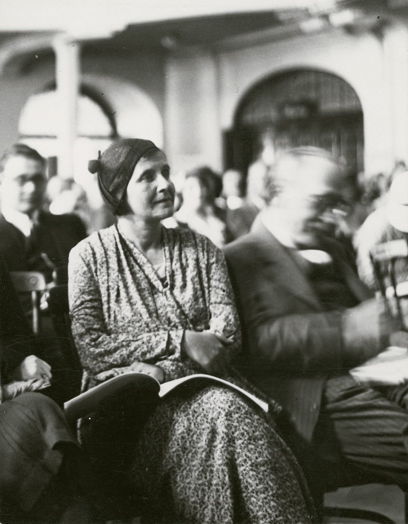Historisches Schwarzweißfoto einer Frau mit einem aufgeschlagenen Buch auf dem Schoss in einem Raum mit vielen Personen.