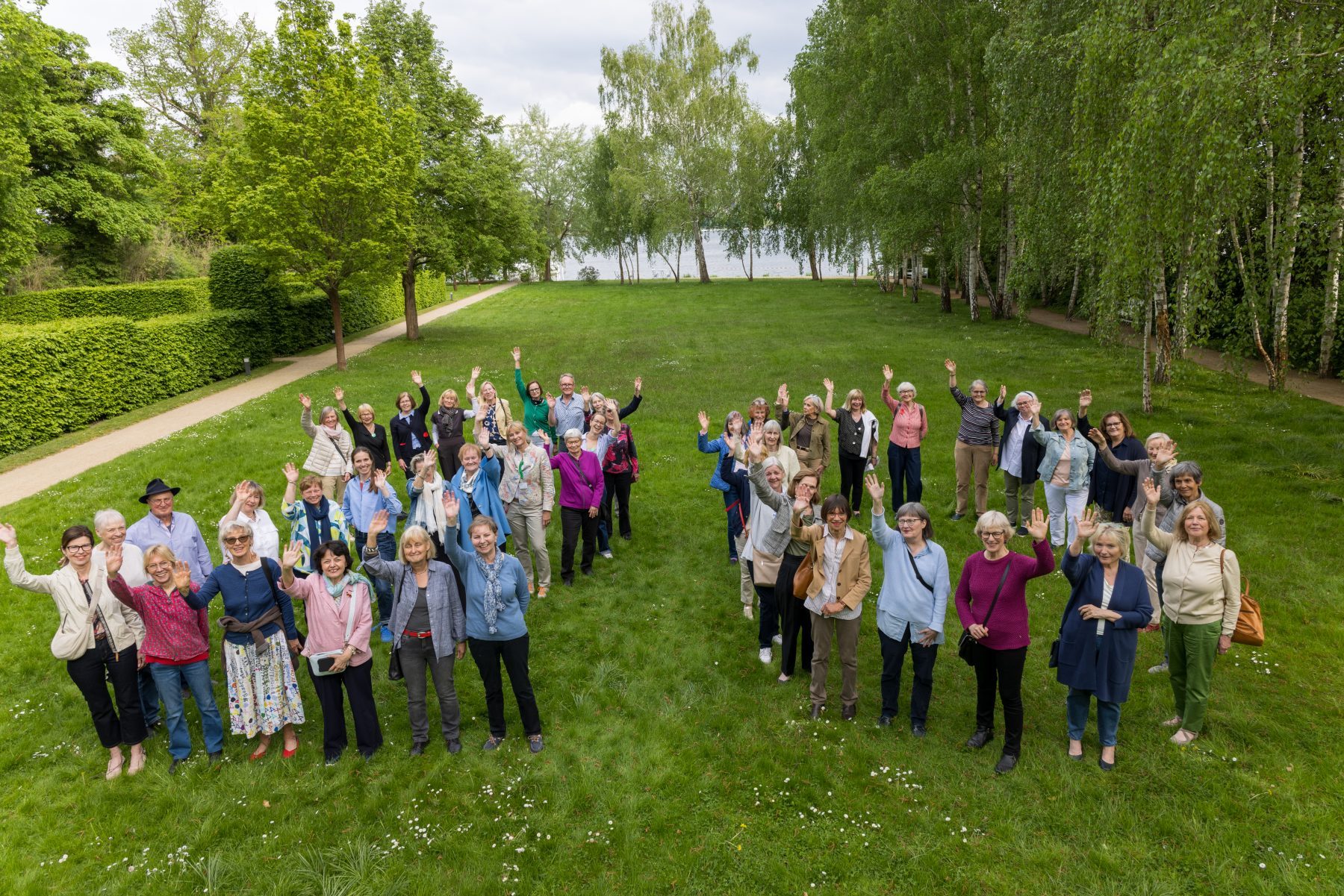 Eine Gruppe bunt gekleideter Menschen formt auf einem Rasen die Zahl 20. Alle winken in die Kamera.