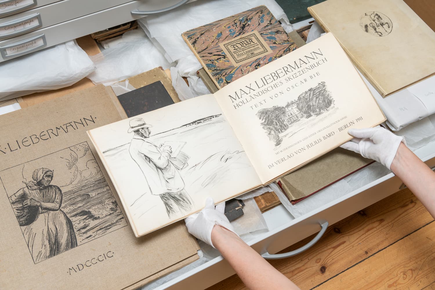 Ein aufgeschlagenes historisches Buch wird über einer geöffneten Schublade mit weiterem historischen Material von zwei Händen mit weißen Baumwollhandschuhen gehalten.
