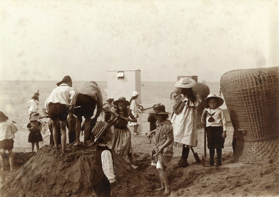 Auf dem historischen Foto spielen Kinder mit Sonnenhüten zwischen Strandkörben im Sand.