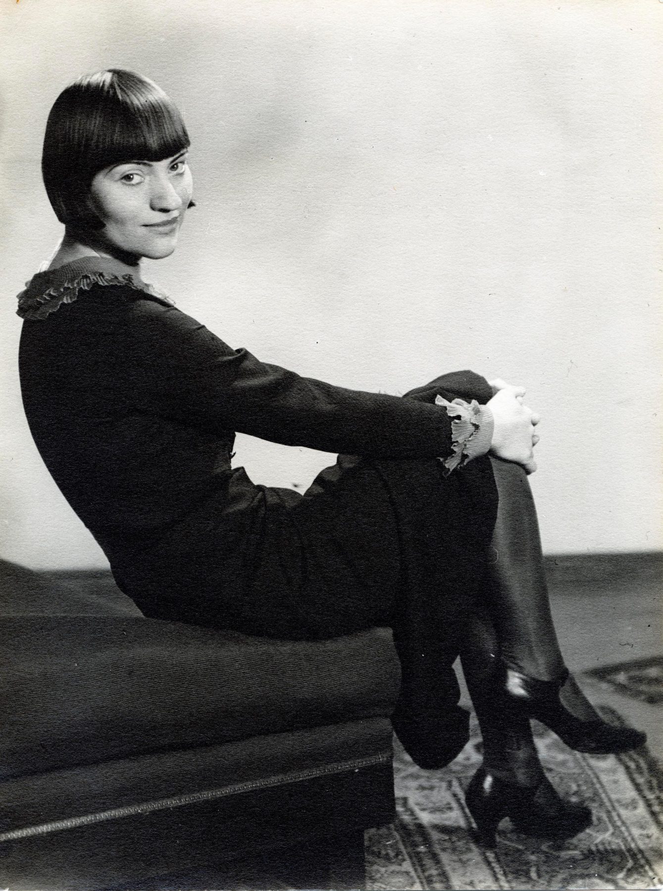Schwarzweißfotografie einer jungen Frau mit Kurzhaarschnitt in einem Kleid, die sitzend und die Beine übereinander geschlage in die Kamera guckt
