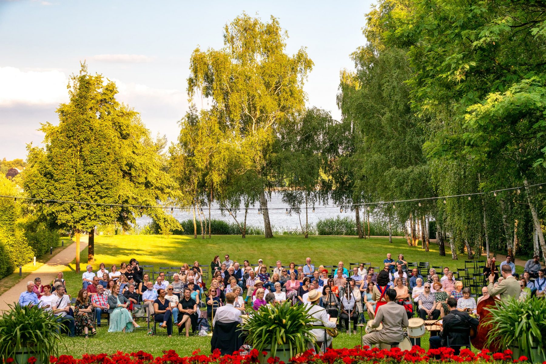 Eine große Gruppe Personen sitzt frontal zur Kamera im Garten der Liebermann-Villa. Im Vordergrund sitzen mehrere Musiker mit dem Rücken zur Kamera.