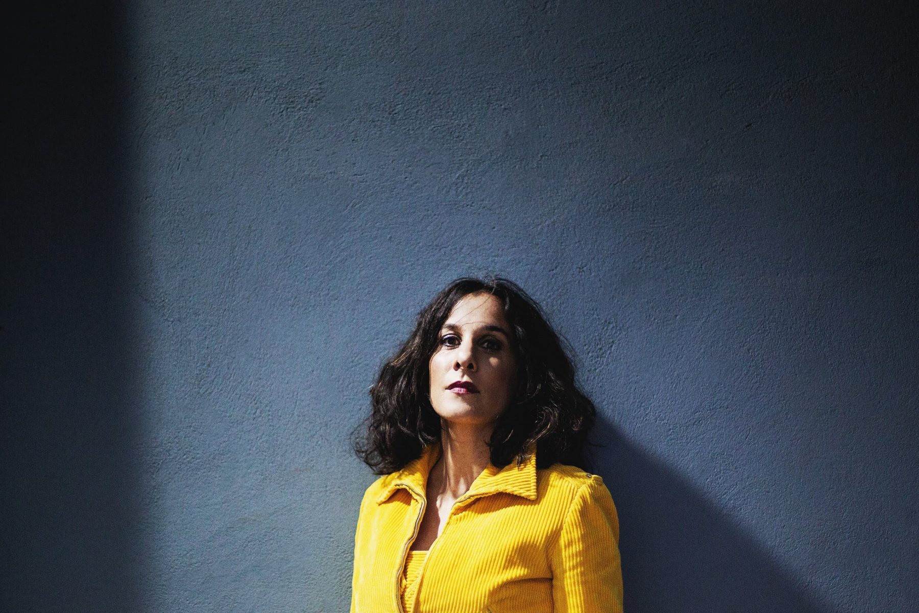 Porträt einer Frau mit schulterlangen dunklen Haaren und einer gelben Jacke vor einer dunkelblauen Wand.