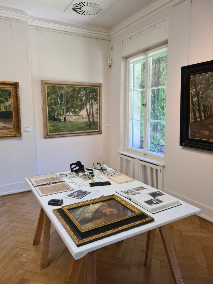 Aktuelle Ansicht eines Ausstellungsraumes mit Tisch in der Mitte, auf dem ein gerahmtes Gemälde sowie Unterlagen und Werkzeuge liegen.