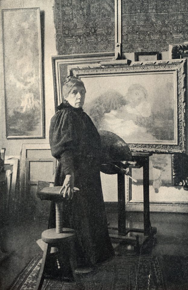 Eine Frau steht vor einer Staffelei auf der ein Gemälde von einer Frau mit Kind steht und hält eine Palette und Pinsel in der Hand.