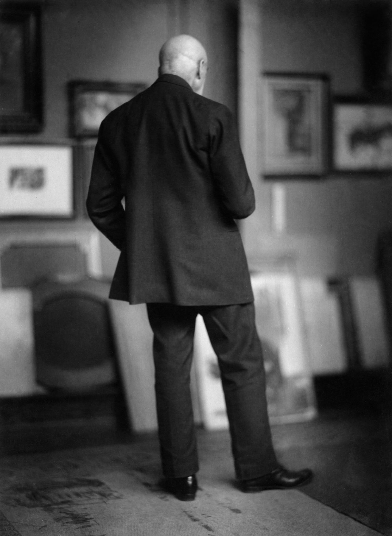 Historisches Schwarzweißfoto einer männlichen Gestalt in einem Anzug von hinten fotografiert in einem Raum mit Kunstwerken