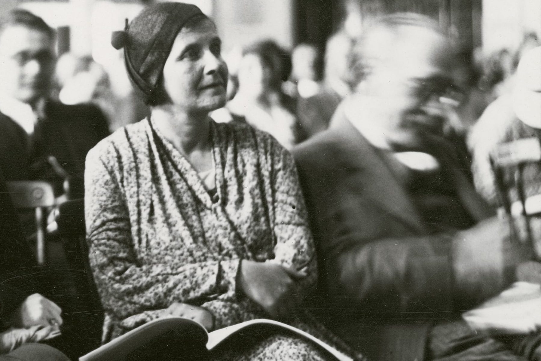 Historisches Schwarzweißfoto einer Frau mit einem aufgeschlagenen Buch auf dem Schoss in einem Raum mit vielen Personen.