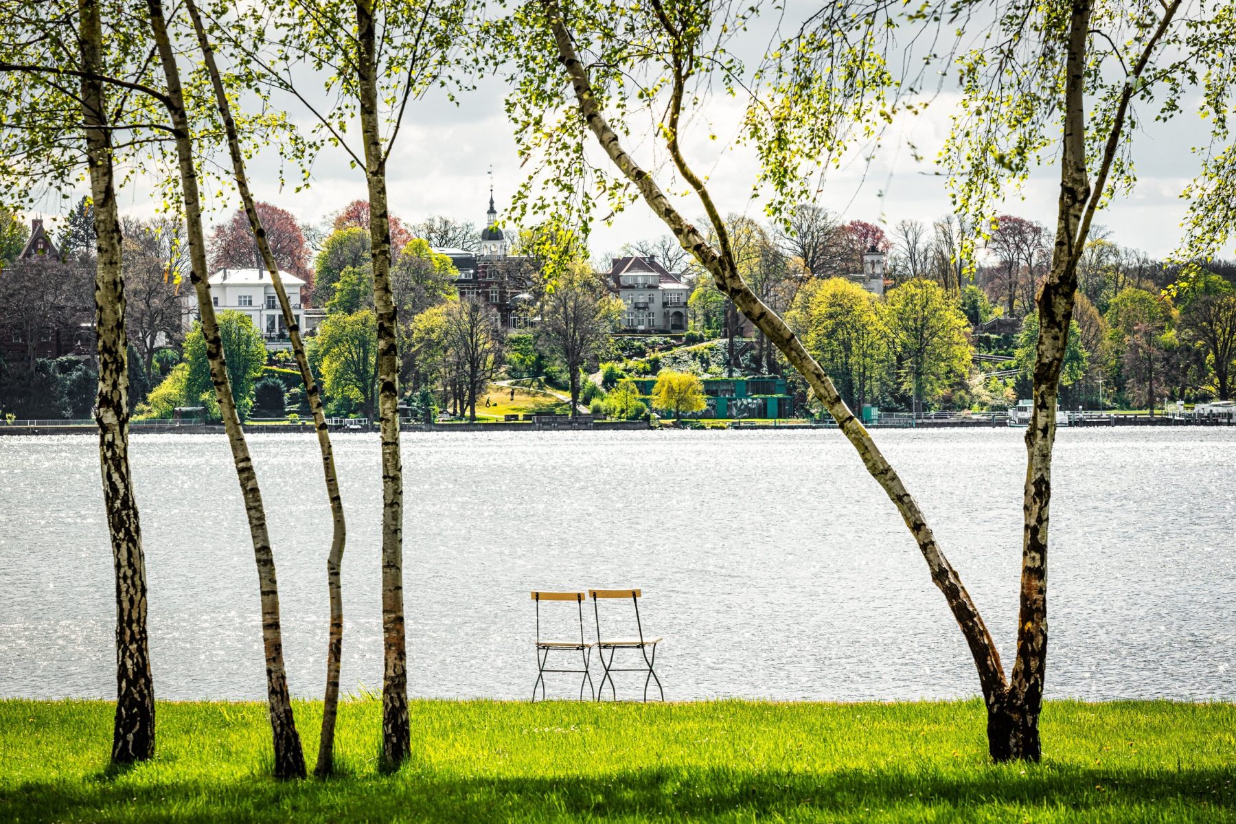 Blick auf das Wannseeufer mit grünem Rasen und zwei Stühlen im Vordergrund und dem Wasser im Hintergrund.