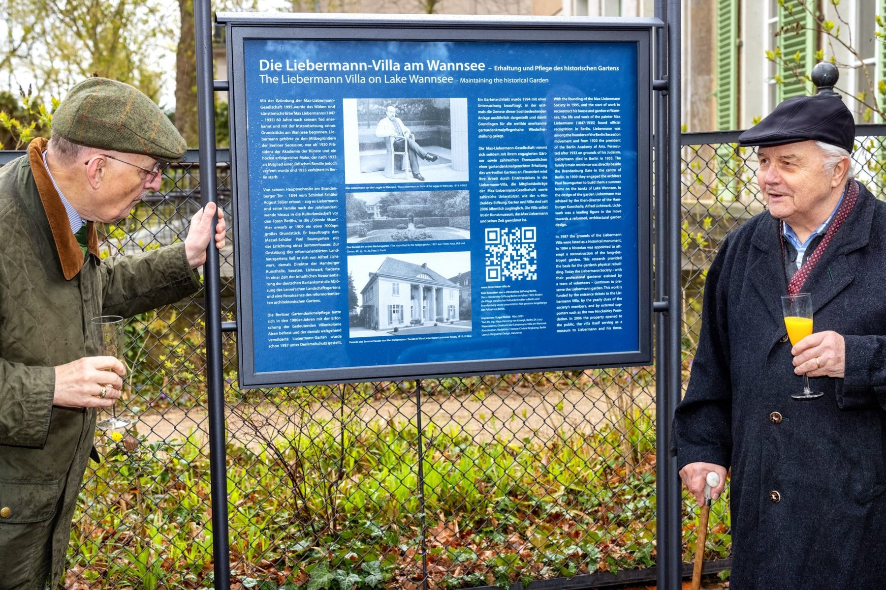 Zwei Männer stehen link und rechts neben einer blauen Infotafel mit Text und Bildern zum Liebermann-Garten.
