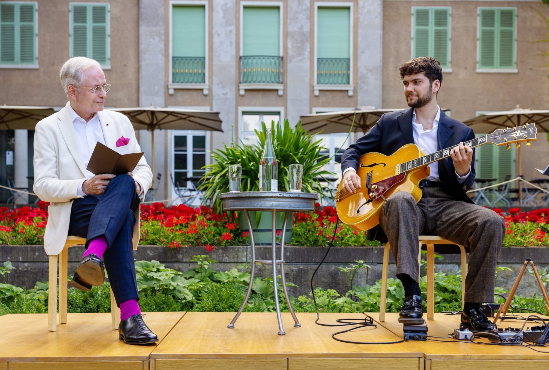 Zwei Persönen auf einer kleinen Bühne in einem Garten, der linke Mann hält ein Buch, der rechte eine Gitarre.