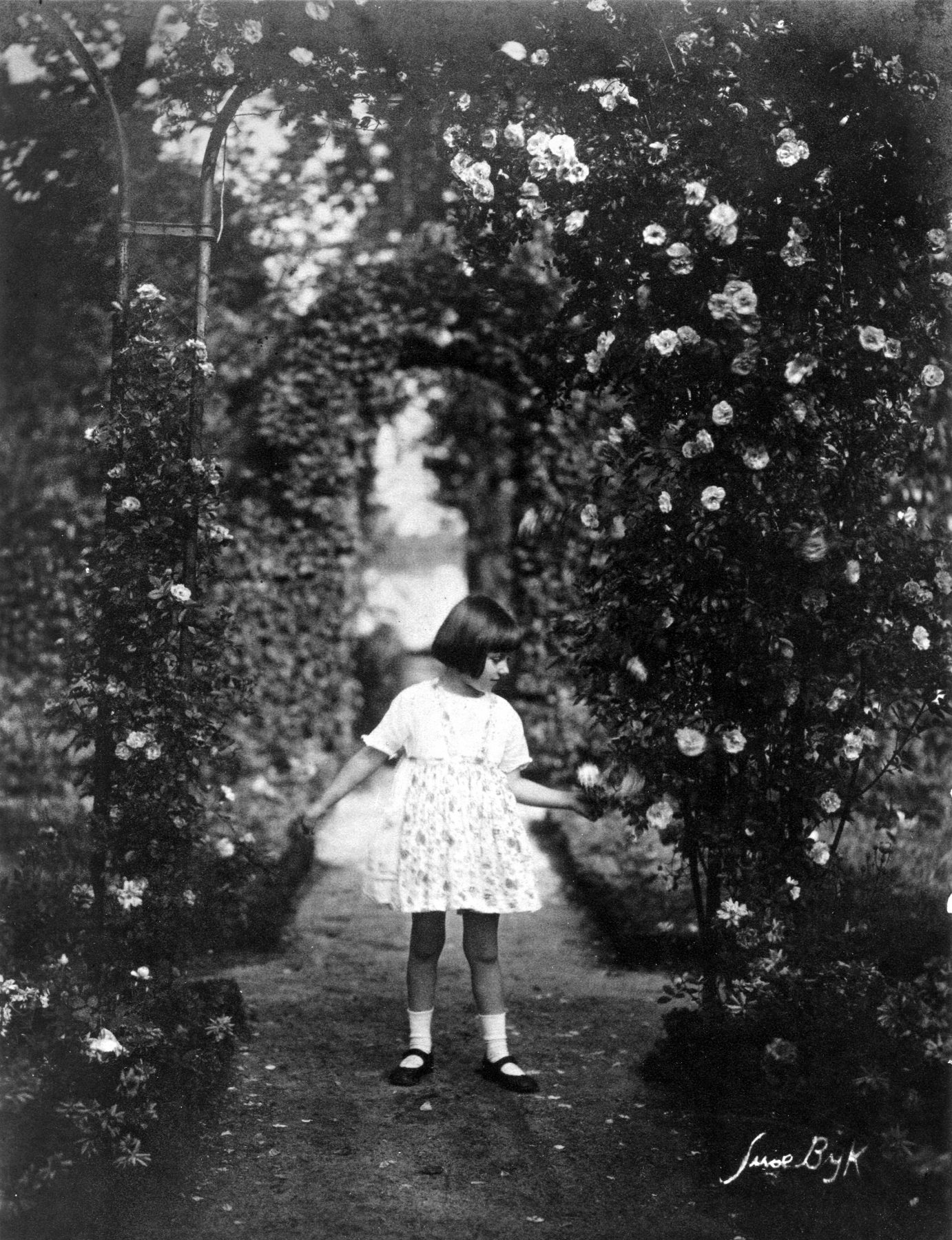 Max Liebermanns Enkelin Maria steht in einem weißen Kleid inmitten der Rosengärten. Der Blick reicht bis zum dahinter liegenden Wannsee.