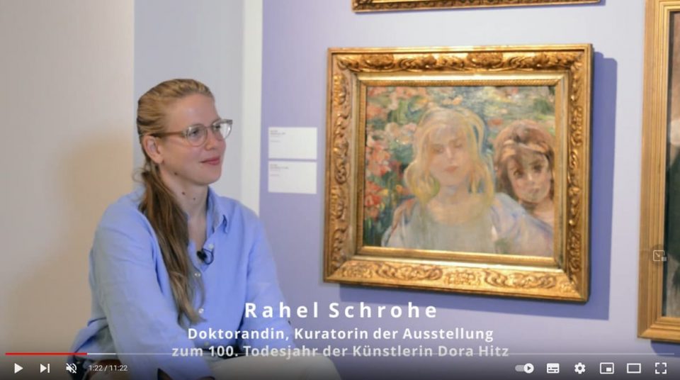 Screenshot eines YouTube-Videos mit einer jungen Frau vor einem Gemälde in einem Ausstellungsraum. Eingeblendeter Text: Rahel Schrohe, Doktorandin, Kuratorin der Ausstellung zum 100. Todestag der Künstlerin Dora Hitz