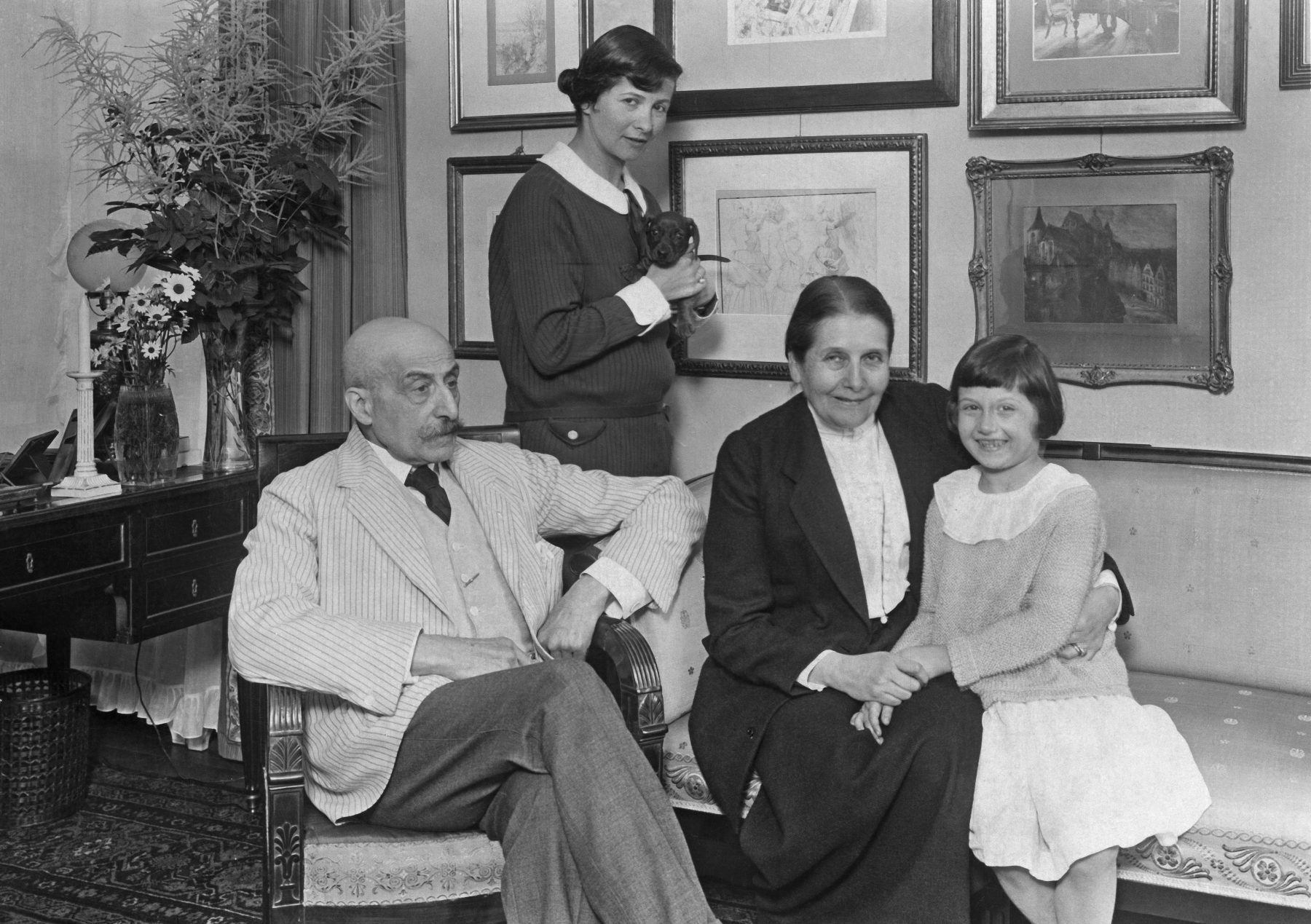 Eine Familie posiert gemeinsam für ein Foto, Links auf einem Sofa sitzt der Künstler Max Liebermann, rechts daneben seine Frau Martha und ihre Enkelin. Dahinter steht die Tochter Käthe mit einem Dackelwelpen.